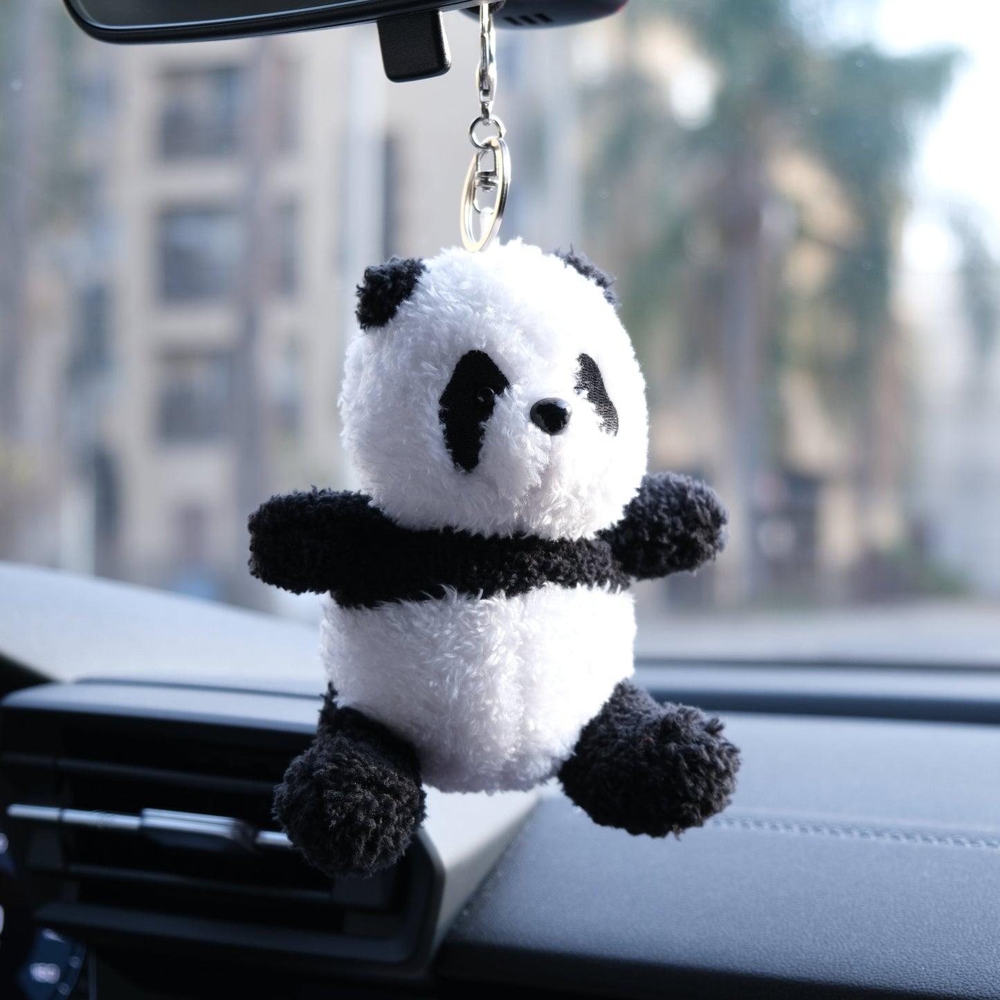 Panda plush keychain hanging from a car's rearview mirror with a blurred outdoor background.
