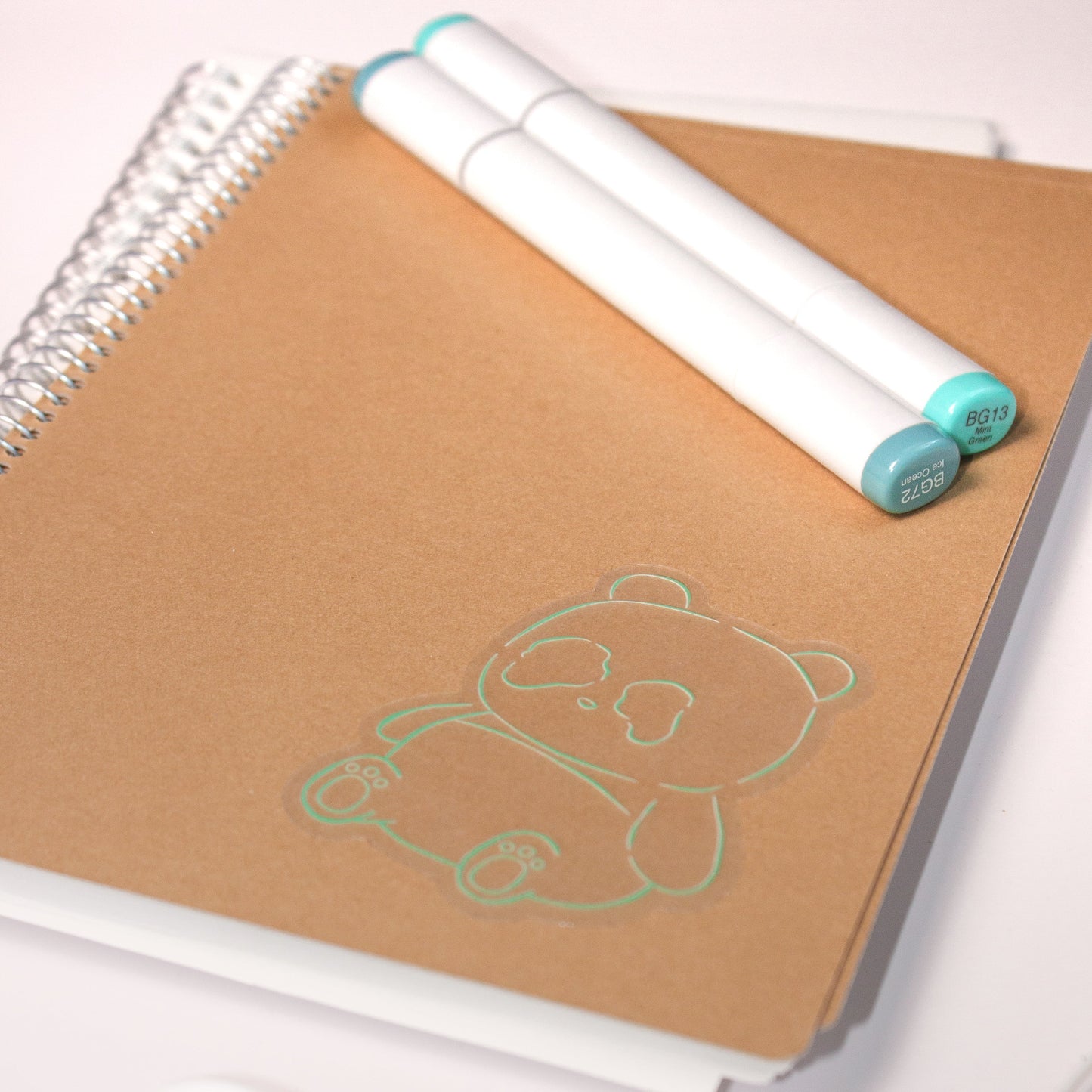 Transparent sticker of panda in a green outline on notebook with a couple of markers